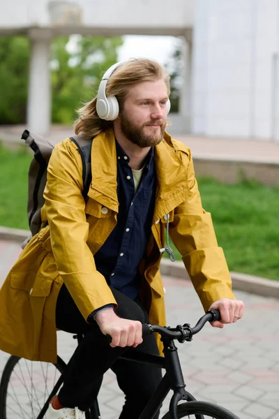 Young bearded man listening to music in wireless headphones while riding a bike along the street