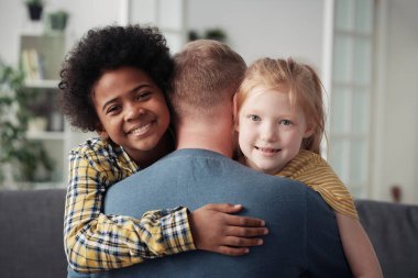 Portrait of happy multiethnic children smiling at camera while embracing their foster dad clipart