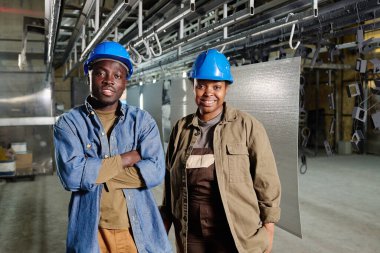 Fabrika atölyesinde durup ekip halinde çalışırken kameraya gülümseyen miğferli Afrikalı meslektaşların portresi