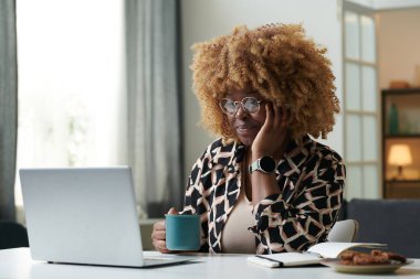 Kıvırcık saçlı Afrikalı genç bir kadın elinde bir fincan kahveyle evde oturuyor ve dizüstü bilgisayarında toplantı yapıyor.