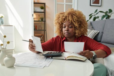 Afrikalı kız oturma odasında oturmuş faturalarını hesaplıyor kira, benzin ve elektrik faturalarını ödüyor.