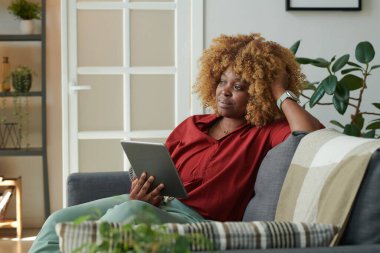 Afrikalı kız oturma odasında kanepede dinleniyor ve dijital tablet kullanırken düşünceli bir ifadeyle kafasını çeviriyor.