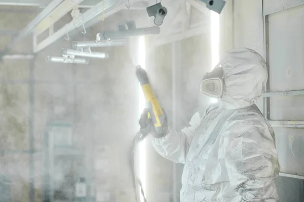 Worker in protective suit working with spray gun to paint metal details at factory