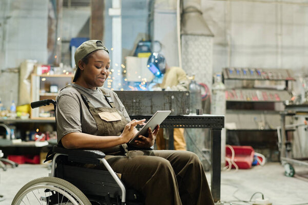 African female worker with disability using digital tablet at work while sitting in wheelchair at workshop
