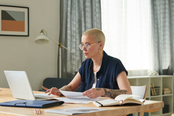 Serious woman in eyeglasses using laptop while working with bills at table in the room