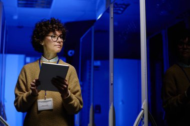 Veri merkezinin koridorunda yürürken tablet kullanan ve sunucu raflarındaki donanımı kontrol eden genç bayan mühendis