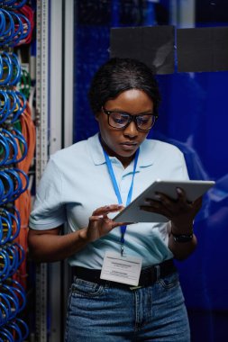 İnternette dijital tablet üzerinde çalışan Afrikalı kadın mühendis, açık rafın yanında internet kablolarıyla duruyor.