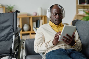 Engelli Afrikalı yetişkin bir adam oturma odasındaki kanepede dinlenmek için dijital tablet kullanıyor.