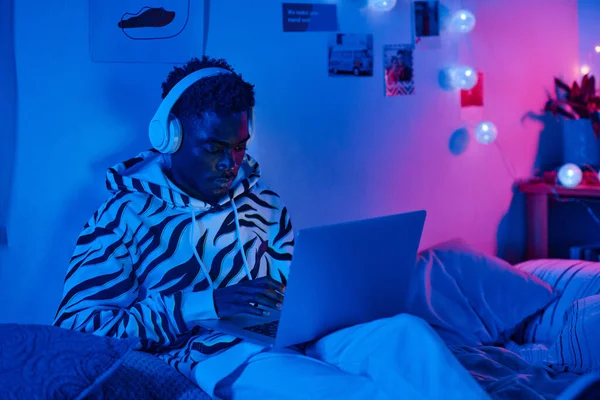 stock image African teenage boy in headphones sitting on bed and playing online game on laptop in dark neon bedroom