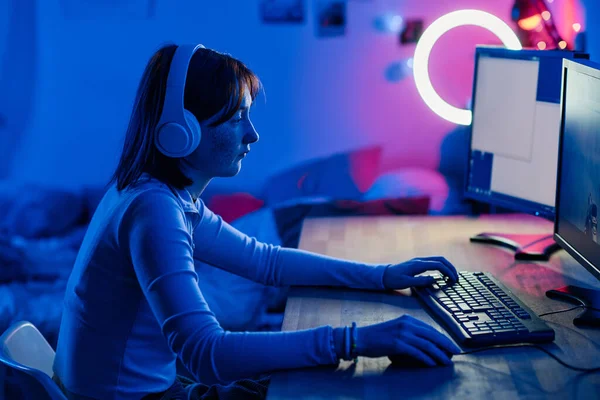 Teenage girl in wireless headphones playing video game on computer while sitting at table in dark neon room