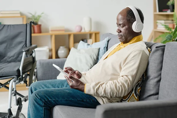アフリカの障害者は ワイヤレスヘッドフォンでソファに座って 部屋で音楽を聴くためにタブレットPcを使用しています — ストック写真