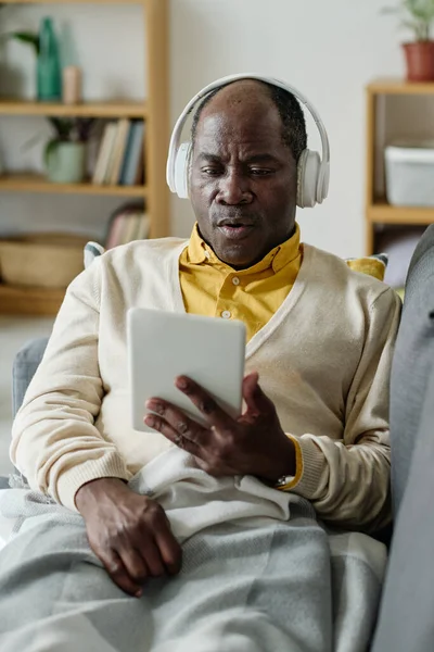 Senior African man in wireless headphones watching video on digital tablet during his rest on sofa at home