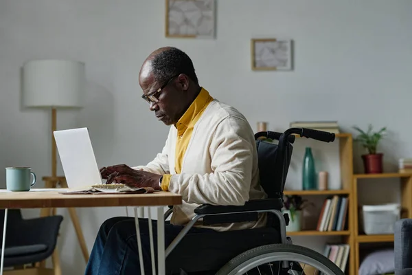 Африканский Мужчина Очках Сидит Инвалидной Коляске Столом Печатает Ноутбуке Дома — стоковое фото
