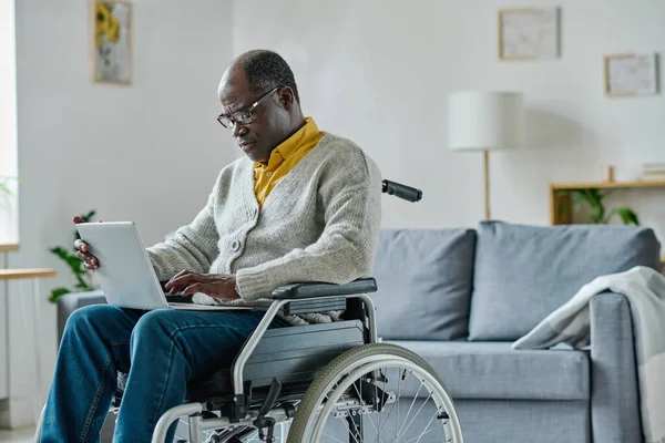 アフリカの成熟した障害のある男性は車椅子に座り 自宅のノートパソコンを使ってオンラインでコミュニケーションをとる — ストック写真