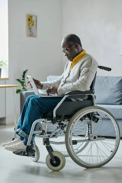 アフリカの成熟した障害のある男性は リビングルームの車椅子に座っているラップトップでのオンライン作業に集中しています — ストック写真