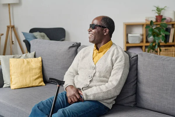 African blind man in dark glasses sitting on sofa in living room and laughing