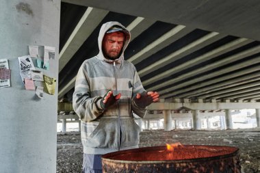 Kirli giysiler içinde evsiz bir adam ellerini ısıtıyor. Açık havada ateşle namlunun önünde duruyor.