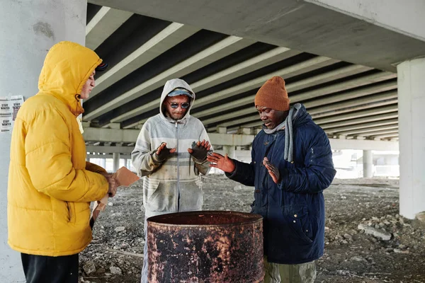 寒い日に屋外で一緒にフェアと暖かいとバレルの近くに立って汚れた服の乞食のグループ — ストック写真