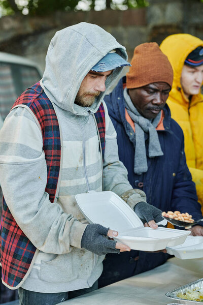 Group of homeless people in warm clothing standing with plastic plates and waiting for food giving by volunteers