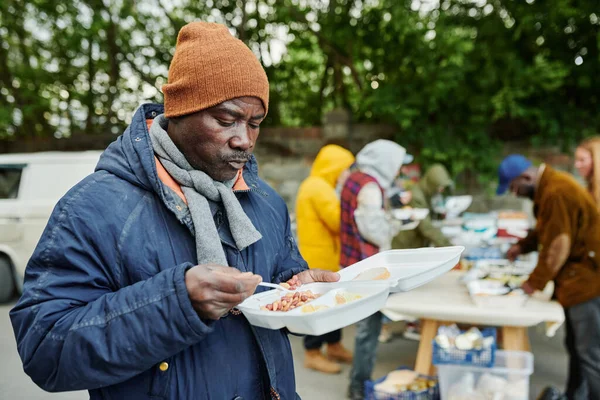 ホームレスのために屋外でボランティアが提供する食べ物を食べる暖かい服のアフリカの貧しい男 — ストック写真