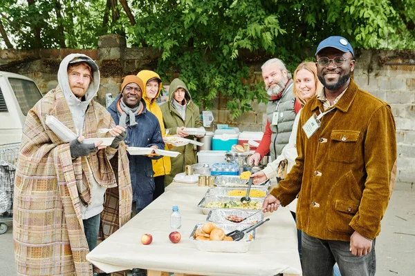 乞食のための慈善事業を配置しながら ボランティアやホームレスの人々のグループの肖像画カメラで笑顔 — ストック写真