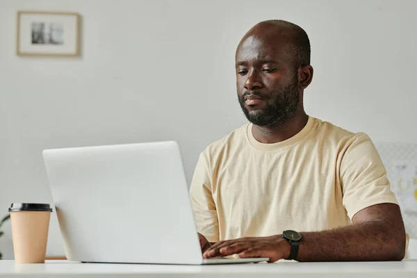 アフリカの若いビジネスマンは テーブルに座ってノートパソコンを入力し 彼は現代のオフィスでオンラインで働いています — ストック写真