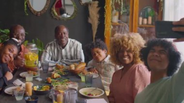 Büyük bir Afro-Amerikan ailesi yemek masasında oturmuş, gülümsüyor ve akıllı telefon kamerasına poz veriyorlar.