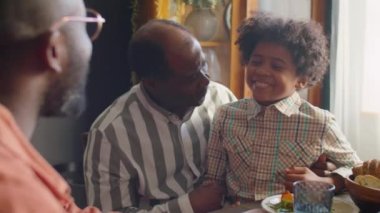 Neşeli Afrikalı Amerikalı çocuk babasının dizlerinin üstüne çöküp aile üyeleriyle akşam yemeği partisinde sohbet ediyor.