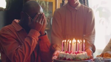 Şaşırmış Afrikalı Amerikalı adam elleriyle gözlerini kapatıyor, sonra doğum günü pastasına mum üflüyor ve ailesiyle el çırpıyor.