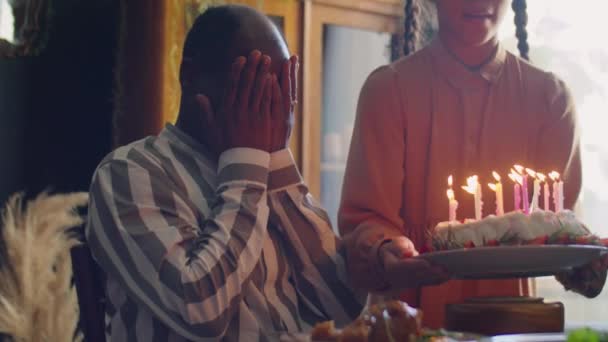 陽気なアフリカ系アメリカ人の男は手で目を覆い 娘は誕生日ケーキをキャンドルで持ち込み その後 家族と一緒に家のお祝いで女の子に抱擁を与えます — ストック動画