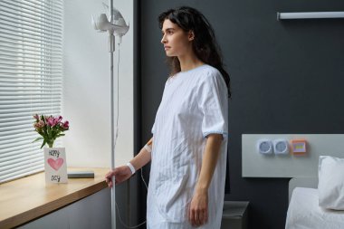 Gömlekli genç bir kadın damlatıcıyla koğuşta dikilirken pencereden dışarı bakıyor.