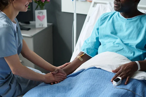 Close-up of young nurse holding hands with elderly patient and talking to him while he sitting on bed with dropper and pulse oximeter