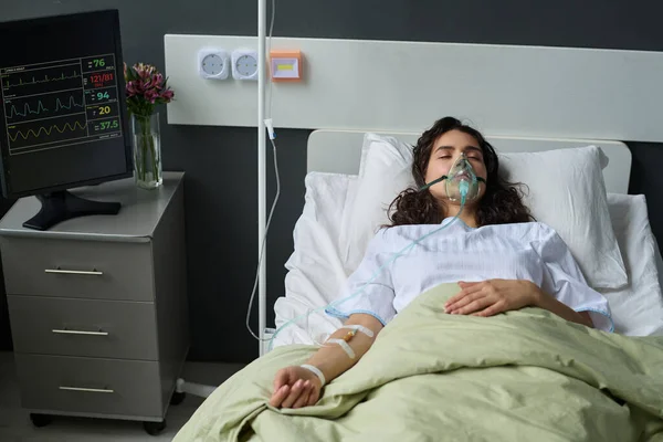 Unconscious young woman lying on bed with oxygen mask during treatment in hospital ward