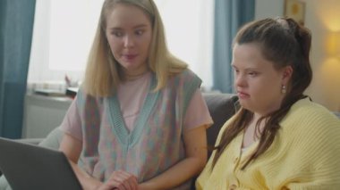 Evdeki kanepede yan yana otururken Down sendromlu bir kıza dizüstü bilgisayar kullanmayı öğreten genç bir kadın.
