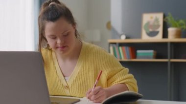 Down sendromlu bir kız evde masa başında otururken dizüstü bilgisayardan not alıyor ve internette geziniyor.