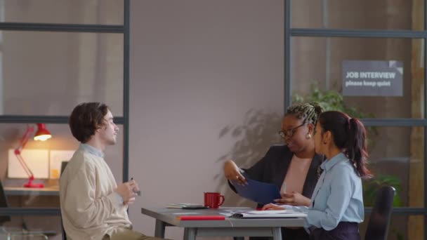 若い白人男性がオフィスに入り 机に座り 履歴書を提出し 仕事の面接中にヒスパニック系とアフリカ系アメリカ人の女性のリクルーターと話します — ストック動画