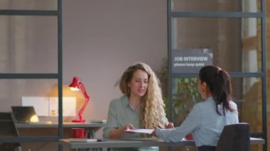 Beyaz iş kadını ofise giriyor, işe alan kişiye özgeçmiş veriyor ve iş görüşmesi sırasında onunla konuşuyor.