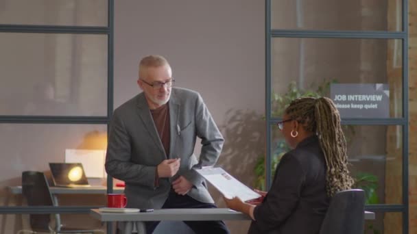 シニア白人ビジネスマンはオフィスに入り アフリカ系アメリカ人女性のリクルーターに履歴書を渡し 面接中に彼女と話す — ストック動画