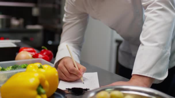 レストランキッチンでの作業中に食品成分のリストを書いてシェフのショットを傾ける — ストック動画