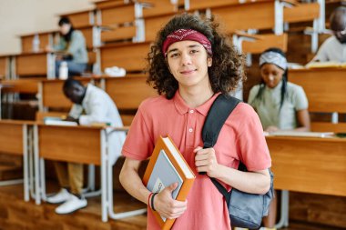 Sırt çantalı ve kitaplı bir öğrencinin portresi, arka planda sınıf arkadaşlarıyla üniversitenin konferans salonunda kameraya bakarken görülüyor.