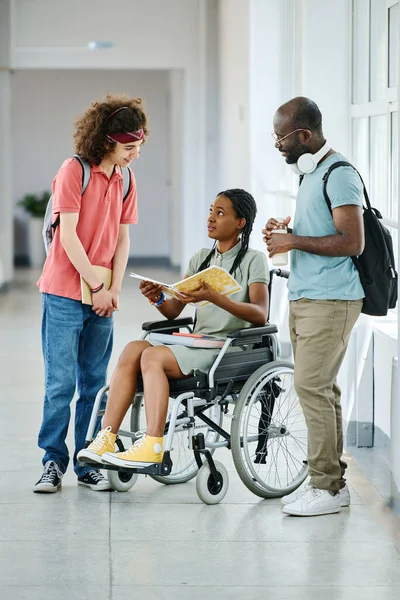 African girl with disability sitting on wheelchair with opened book and discussing homework with her classmates while they standing at corridor