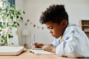 Afrikalı ilkokul çocuğu kitap ve defterle masada oturuyor ve evde ödev yapıyor.