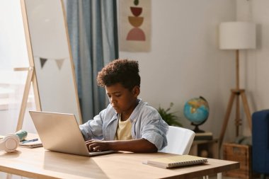 Afrikalı çocuk, odasında otururken ödevlerini internette yapmak için dizüstü bilgisayarda yazı yazıyor.