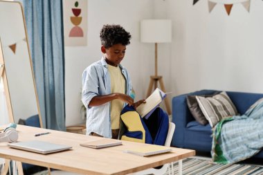 Afrikalı okul çocuğu sırt çantasına kitap koyuyor. Odasında duruyor, okula hazırlanıyor.
