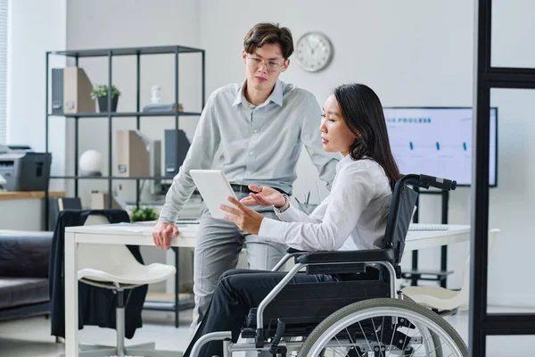 Asian businesswoman with disability pointing at digital tablet and discussing online report with her colleague at office