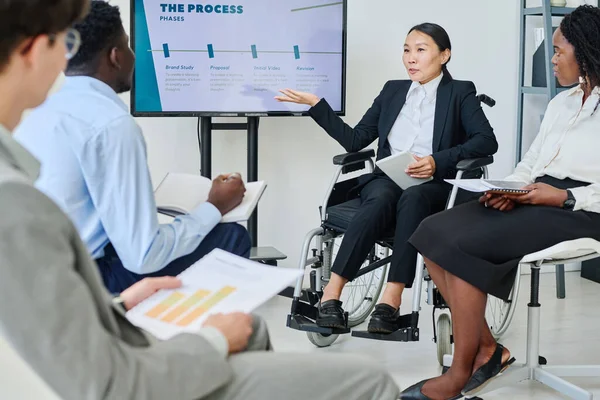 Asian business leader with disability presenting her report to colleagues at presentation, she sitting on wheelchair and pointing at the screen