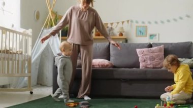 Küçük çocuk ilk adımlarını atarken kardeşinin yanına yürüyordu. Annesinin yardımıyla evdeki çocuk odasında.