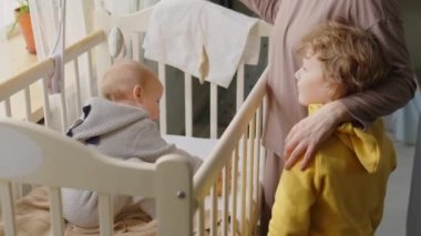 Anne küçük oğlunun kafasını okşuyor, gülümsüyor ve konuşuyor. Evde beşikte bebekle dikiliyordu.