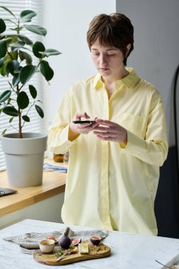 Genç fotoğrafçı masada duruyor ve akıllı telefonunu kullanarak tepsiyi yiyecekle fotoğraflıyor.