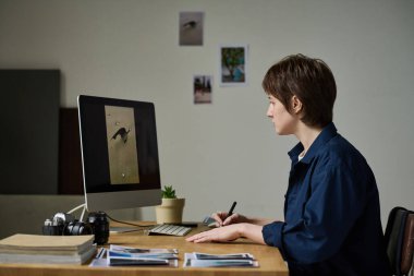 Genç grafik tasarımcısı iş yerinde otururken bilgisayar ekranında profesyonel fotoğrafları rötuşluyor.